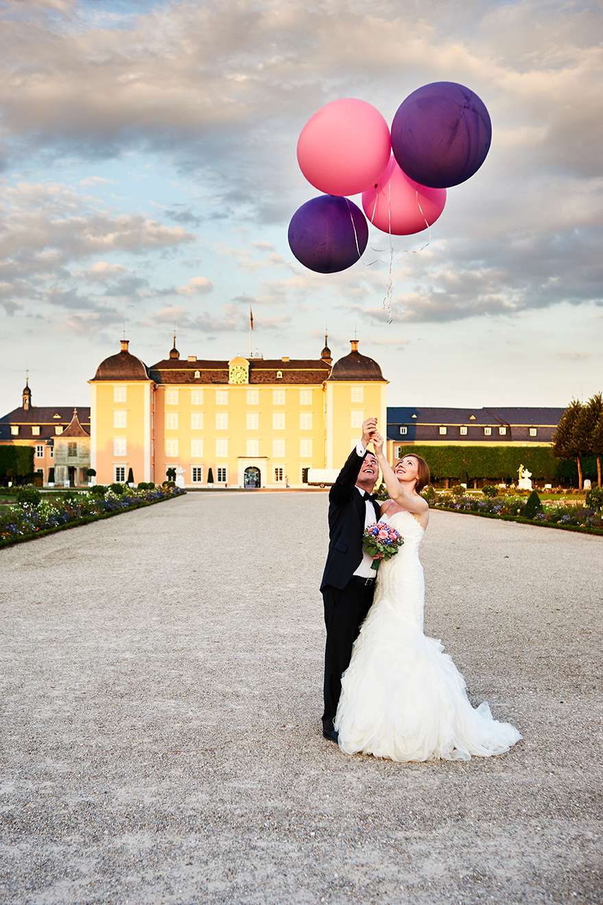 GBrautpaar hält bunte Luftballons, festlicher Moment vor imposanter Kulisse.